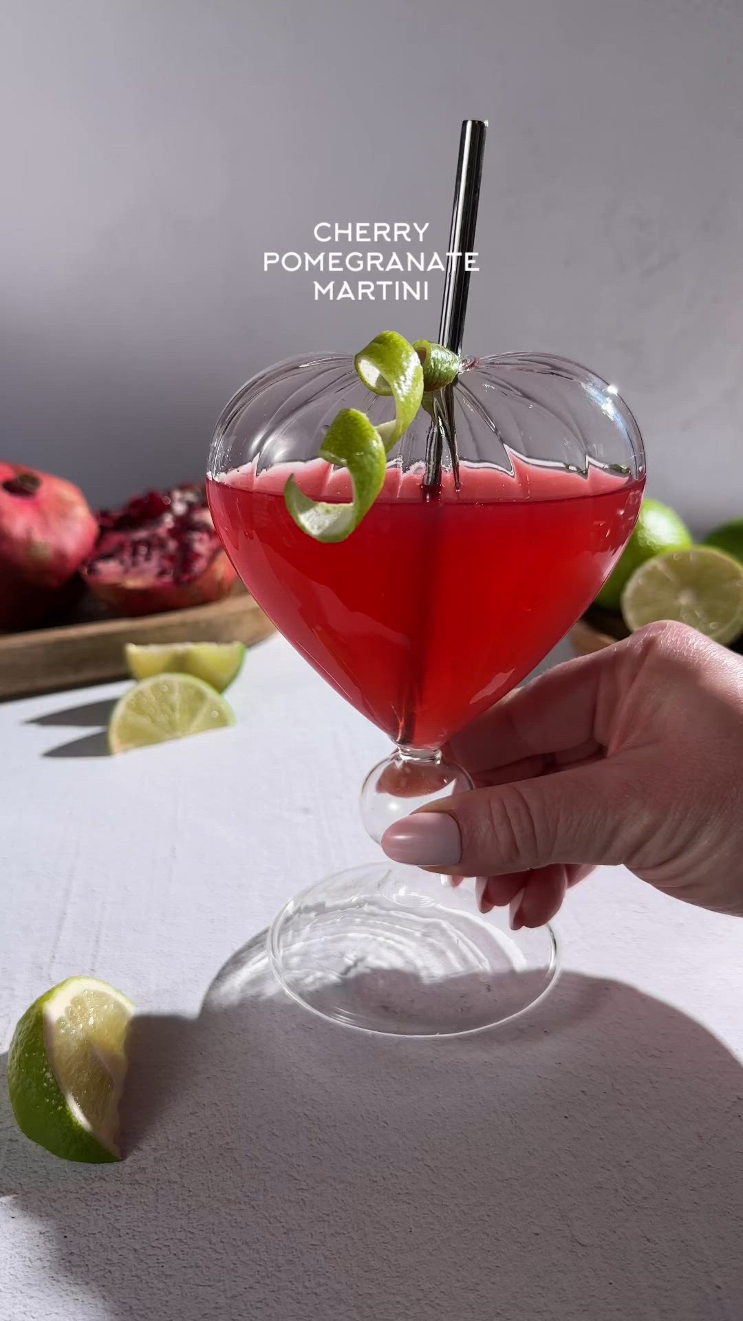 This may contain: a hand holding a martini glass filled with red liquid and garnished with limes