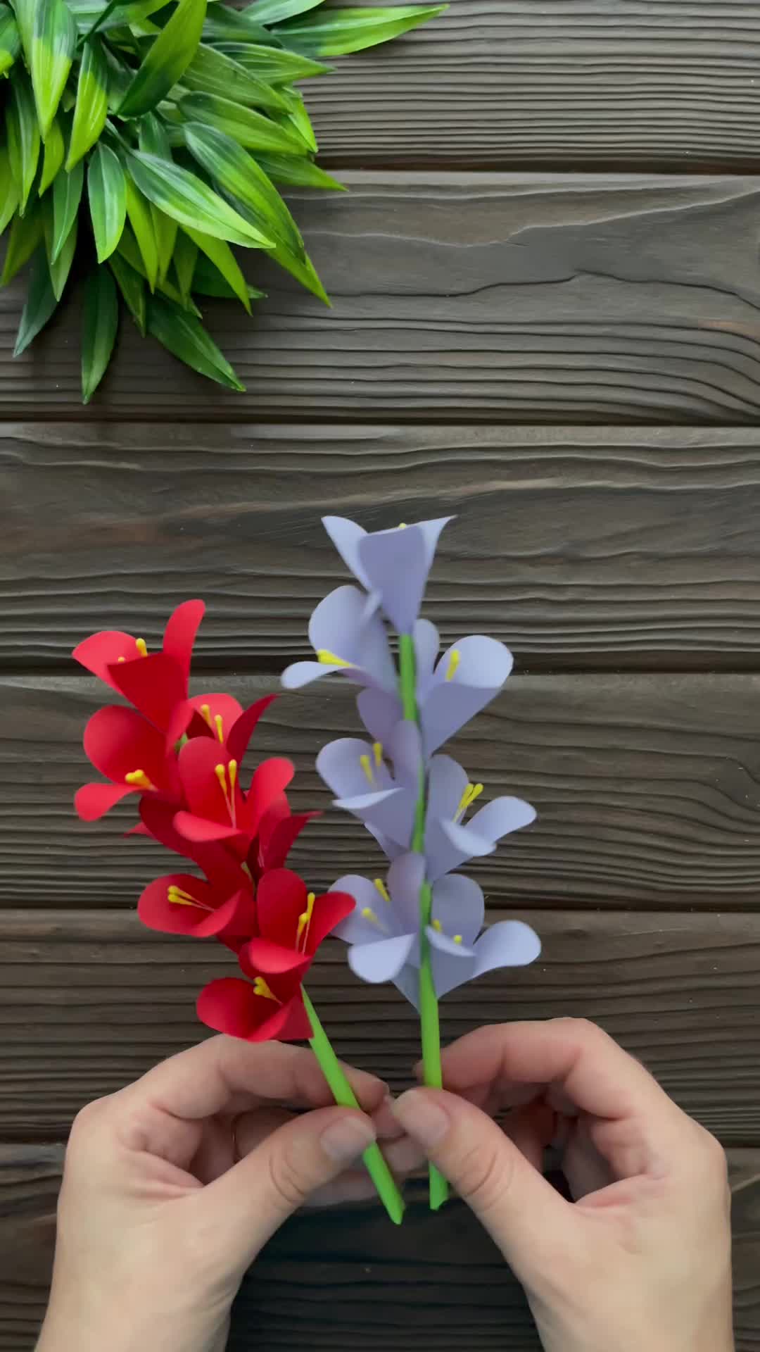 This may contain: a hand is holding some paper flowers in front of a wooden wall and green plants