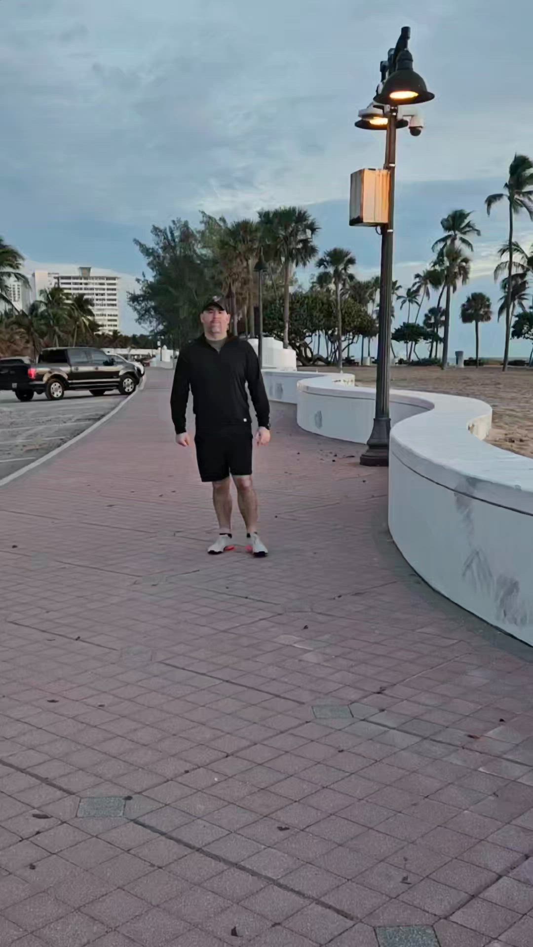 This may contain: a man in black shirt and shorts standing on brick walkway next to street light with palm trees