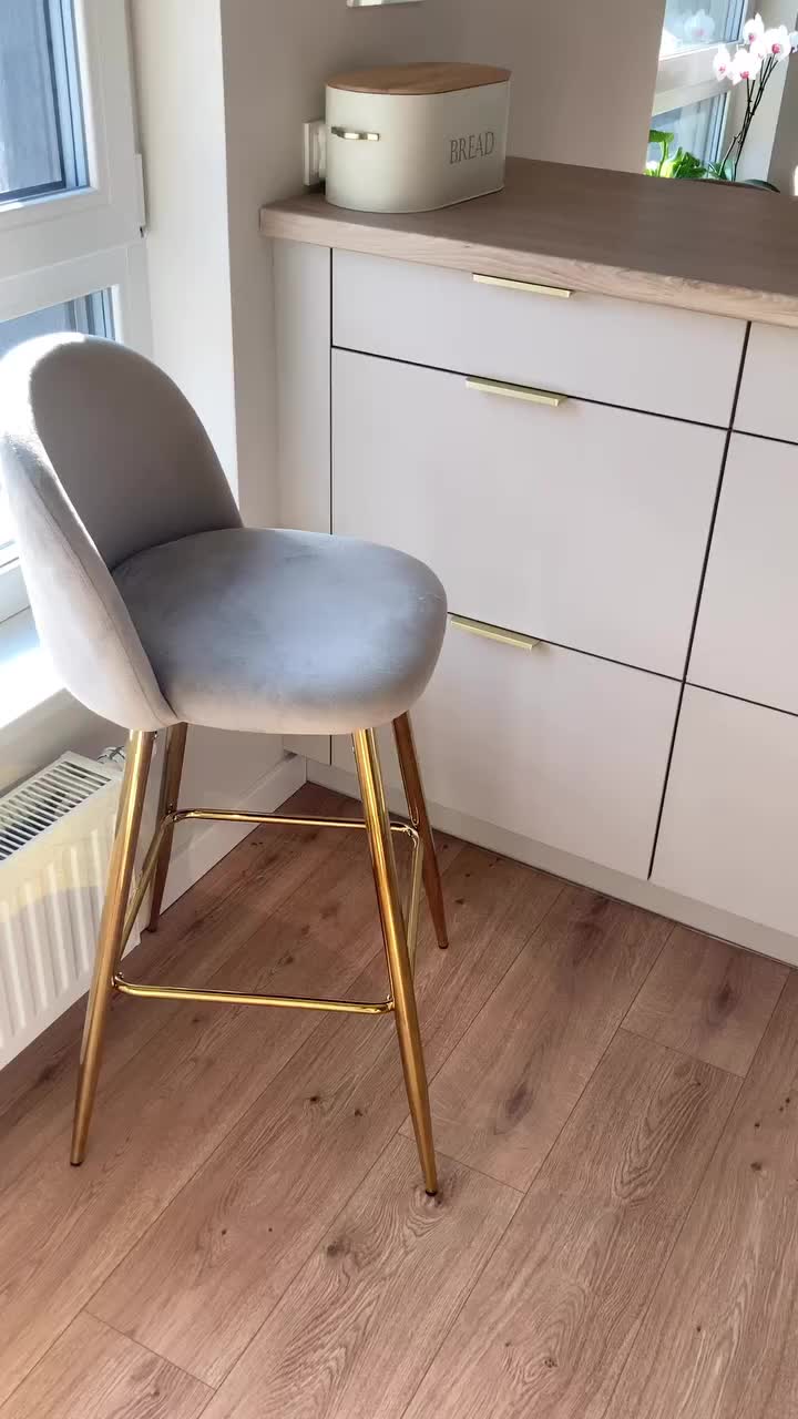 This may contain: a grey and gold bar stool in front of a kitchen window with white cupboards