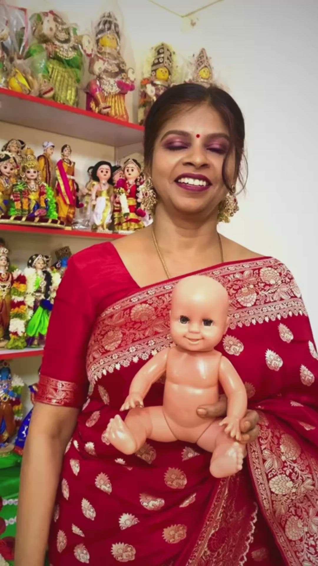 This may contain: a woman in a red sari holding a baby doll and smiling at the camera