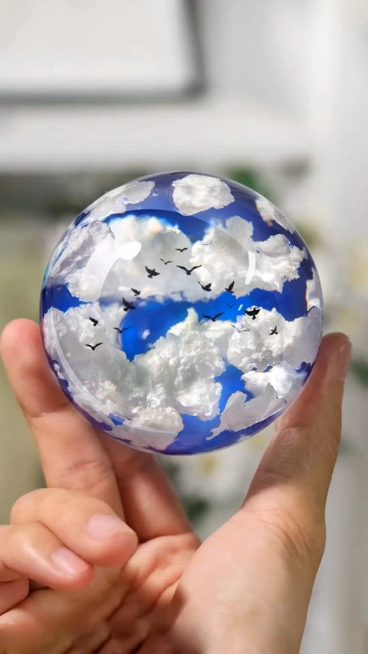 This may contain: a hand holding a glass ball with clouds and birds in the sky painted on it