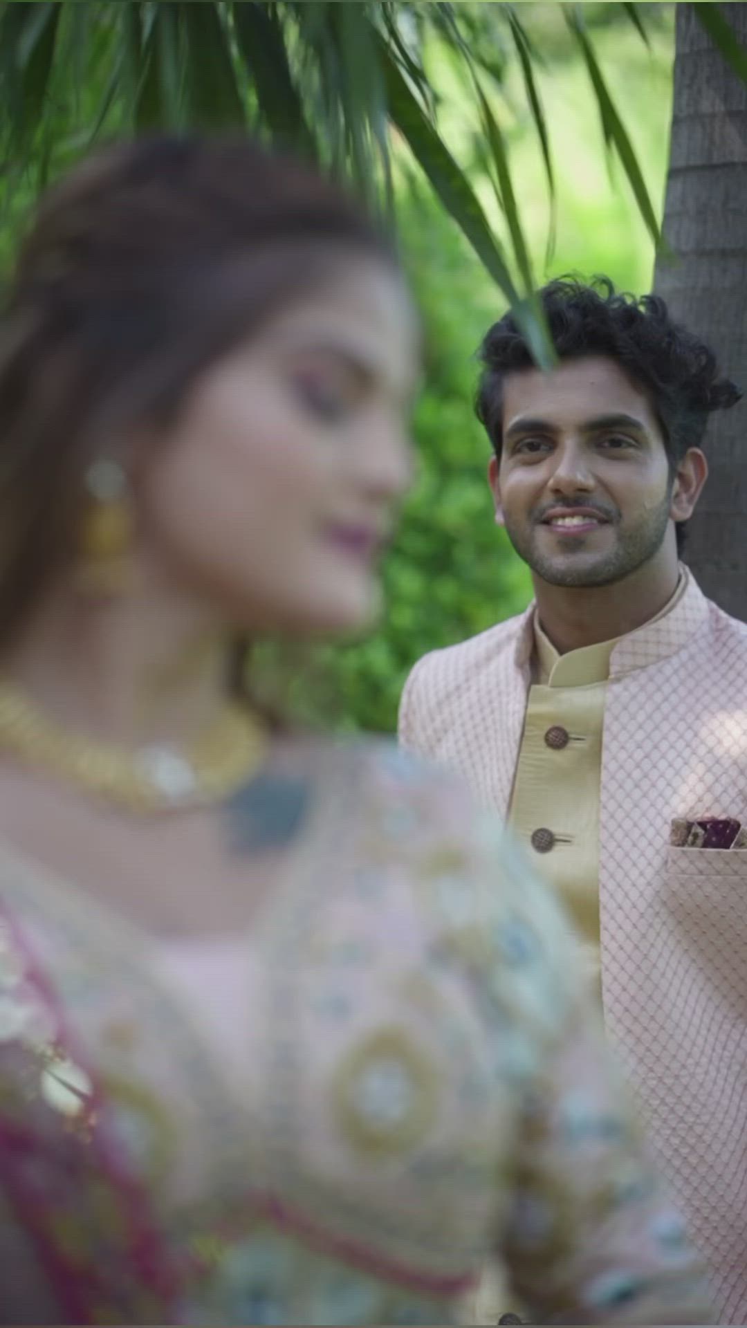 This may contain: a man and woman standing next to each other in front of some trees, one wearing a pink suit