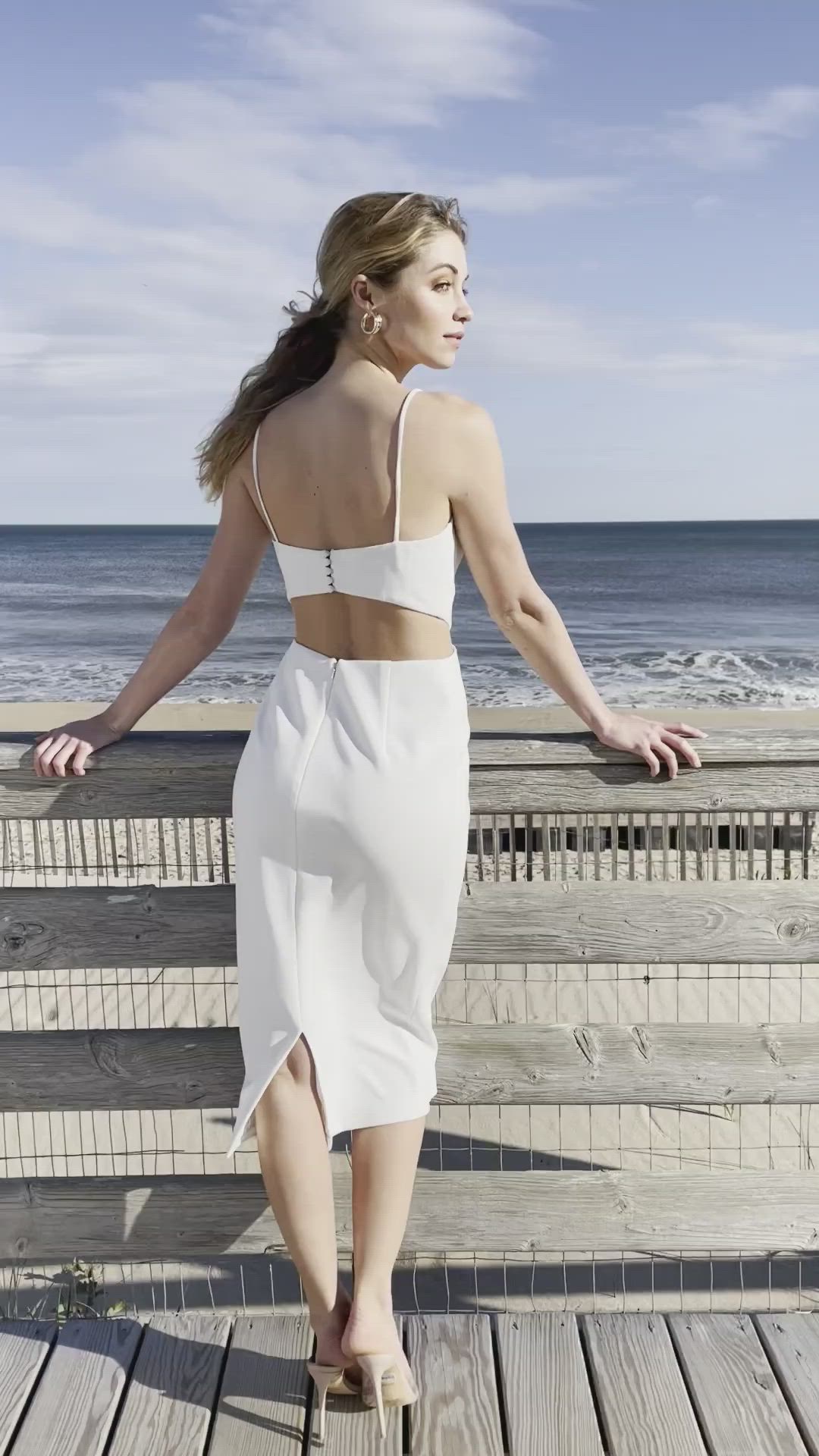 This may contain: a woman in a white dress standing on a wooden deck next to the ocean and holding a purse