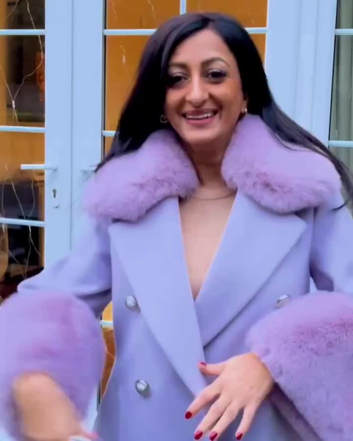 This may contain: a woman with long black hair wearing a purple coat and holding her hand out to the side