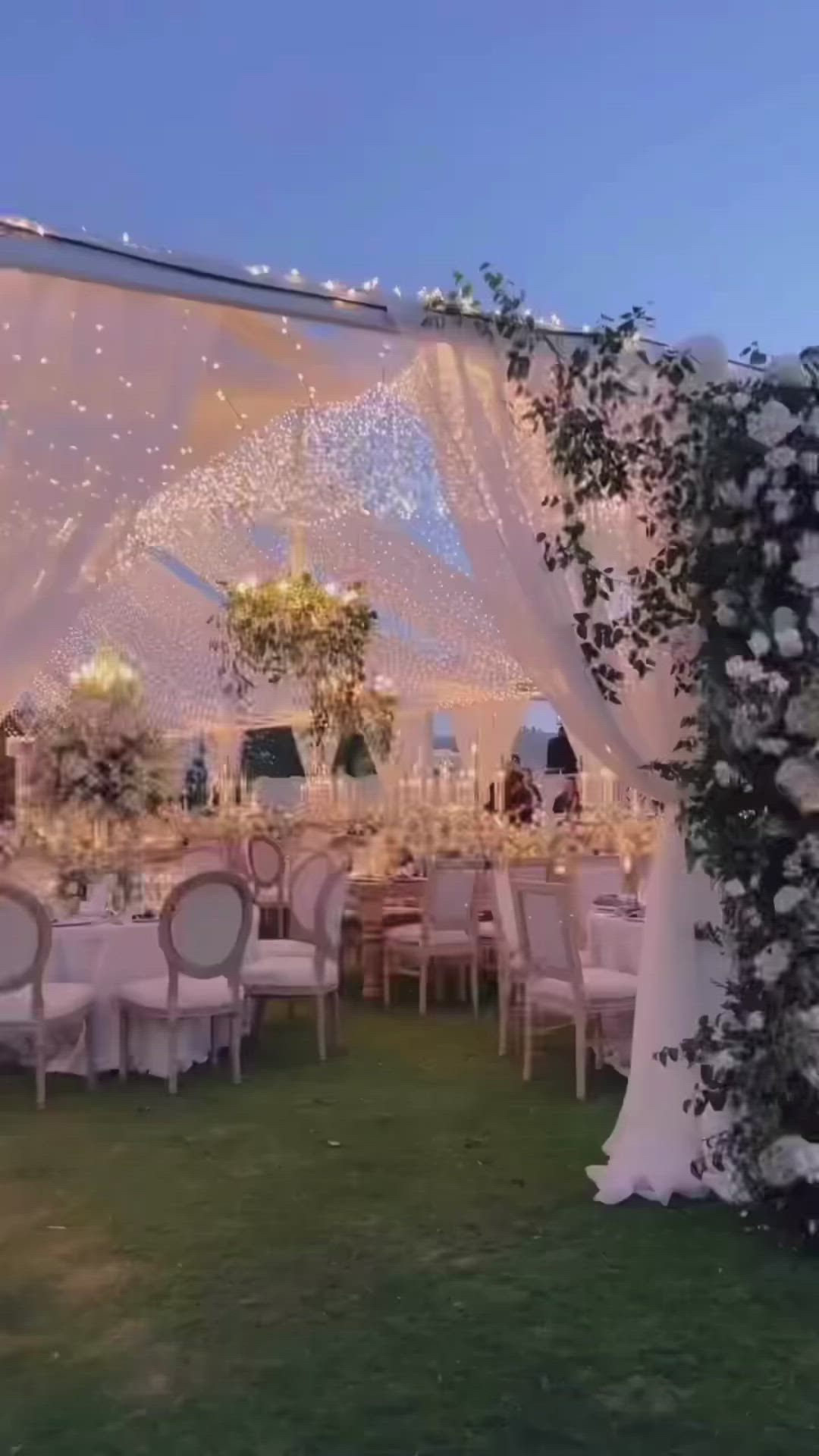 This may contain: an outdoor wedding reception setup with white linens and greenery on the grass, surrounded by string lights