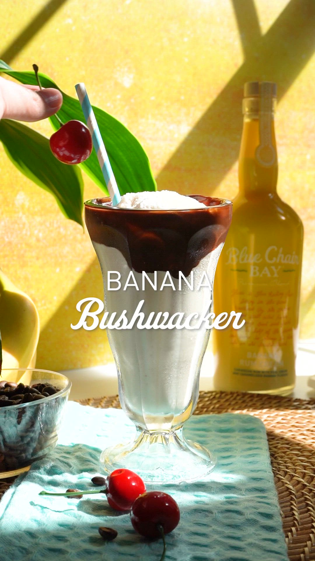 This may contain: a banana bushwaker with cherries in a glass on a table next to two bottles