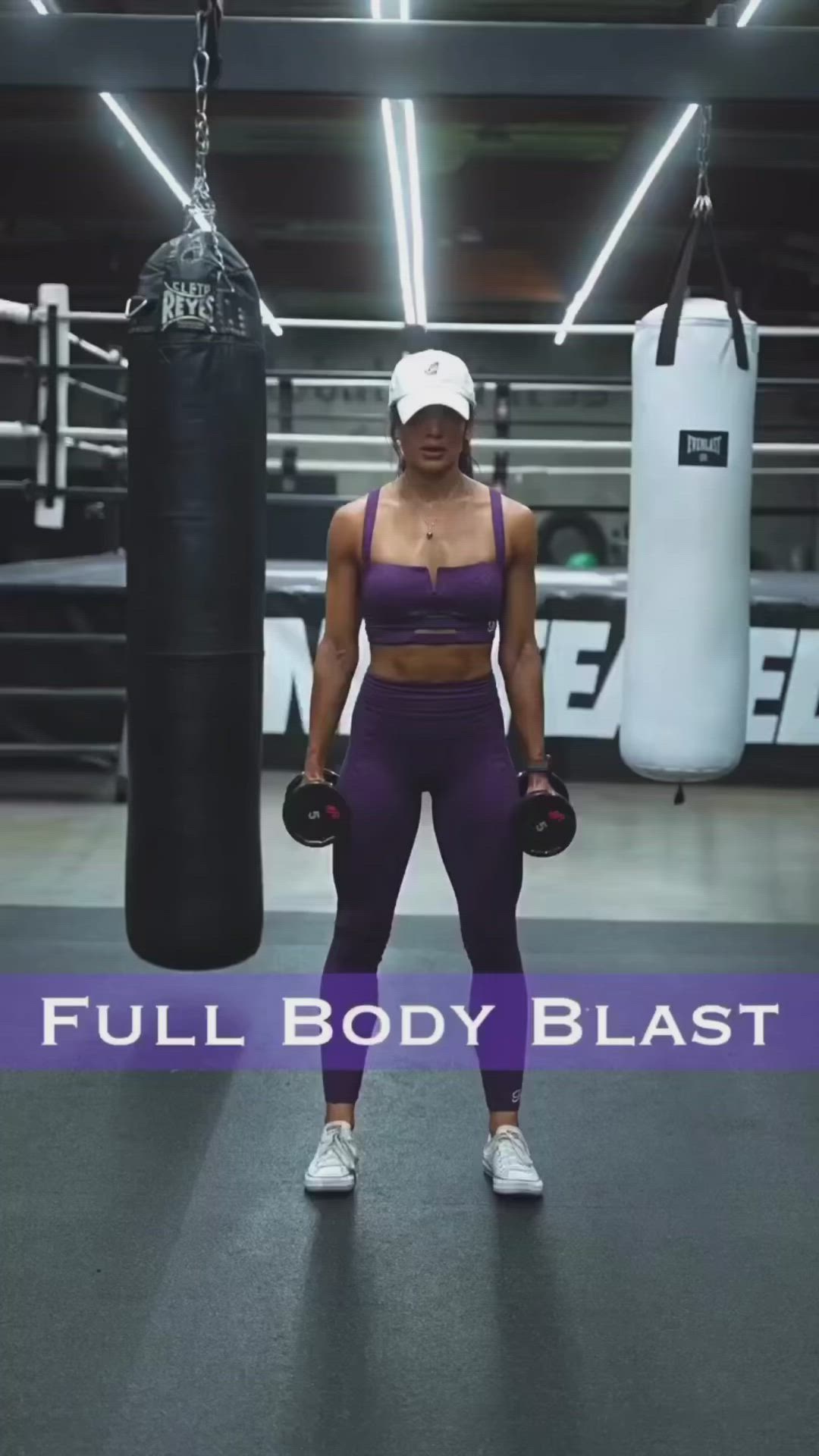 This may contain: a woman in purple tights and white hat is holding a black punching bag with the words full body blast on it