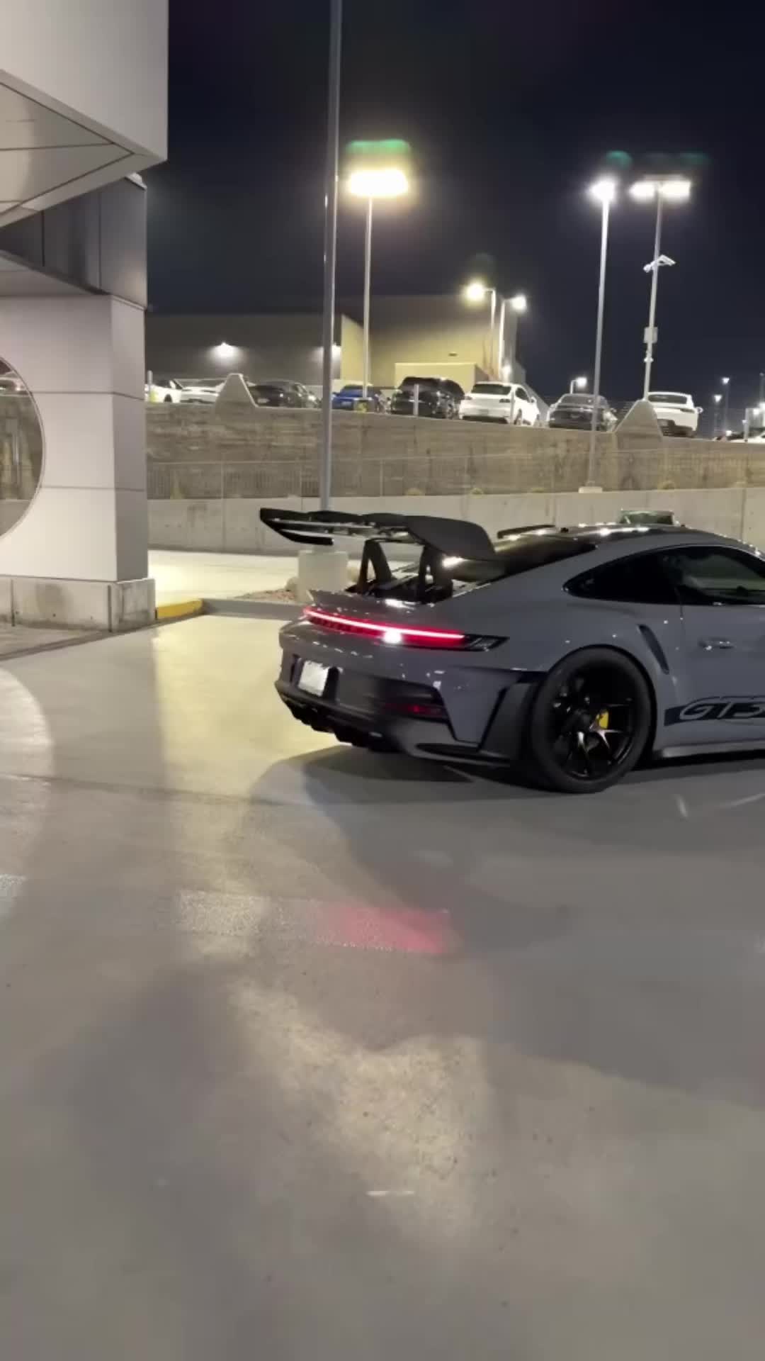 This may contain: a grey sports car parked in front of a parking lot at night with its lights on