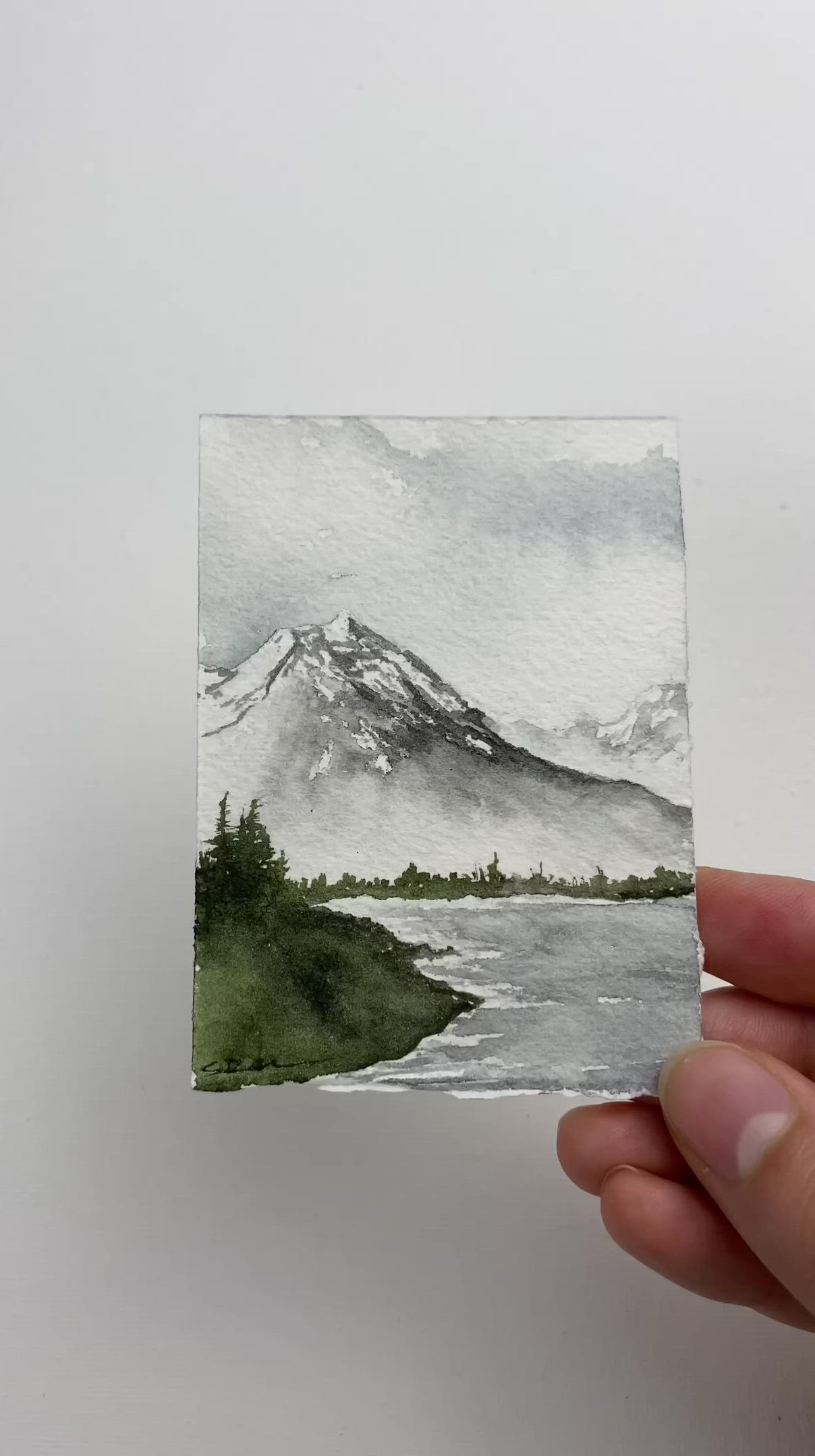 This contains: Miniature watercolor painting process video - snow capped mountains and bright green trees and lake inspiring adventure in small size