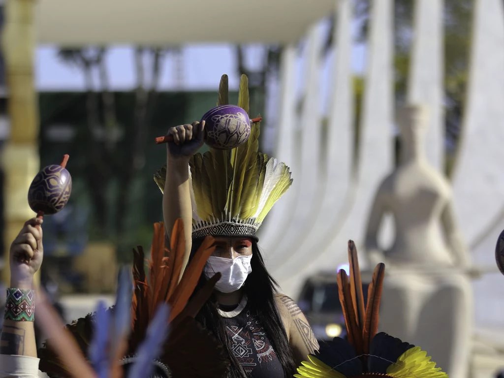 Mulher indígena posa em frente ao STF com punho cerrado e levantado. Ela usa adereços indígenas tradicionais.