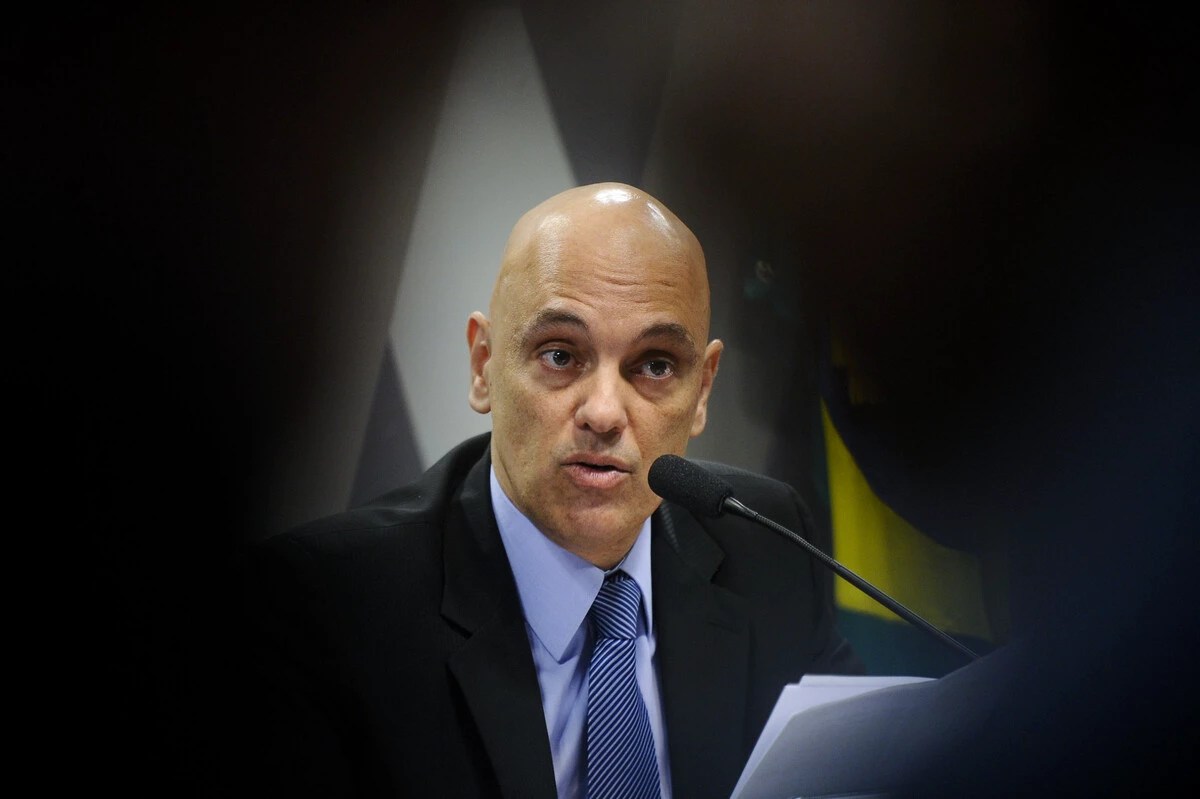 O ministro do STF Alexandre de Moraes é um homem branco, com a cabeça raspada e olhos escuros. Alexandre de Moraes veste paletó preto e gravata azul, ele fala ao microfone.