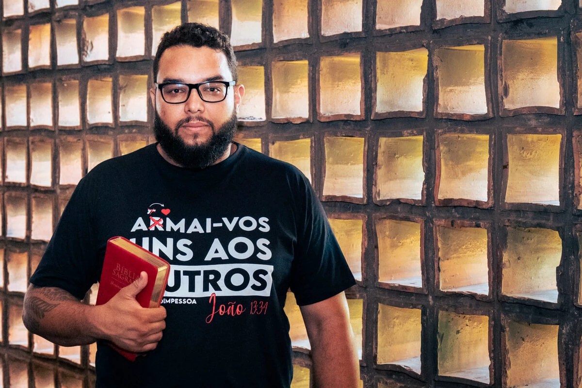 Pastor Filipe Gibran, da igreja Comuna do Reino, em Belo Horizonte (MG). Filipe Gibran é um homem branco com cabelos, olhos e barbas pretos e usa camiseta estampada com versículo bíblico, ele segura em suas mãos uma bíblia vermelha.