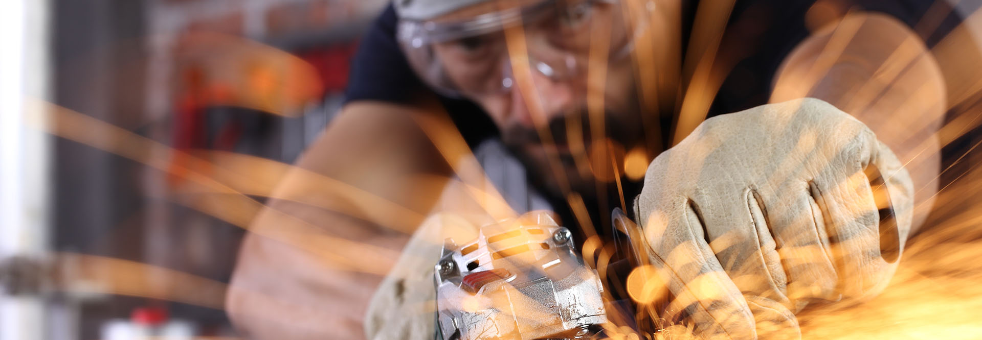 Nahaufnahme von einem Mann, der mit Schutzbrille und Schutzhandschuhen ein Metallwerkstück schleift, das in einem Schraubstock eingespannt ist. Es fliegen orangene Funken.