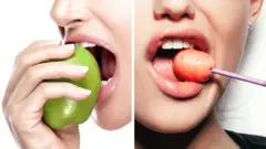 One woman eating an apple, and another eating a lollipop