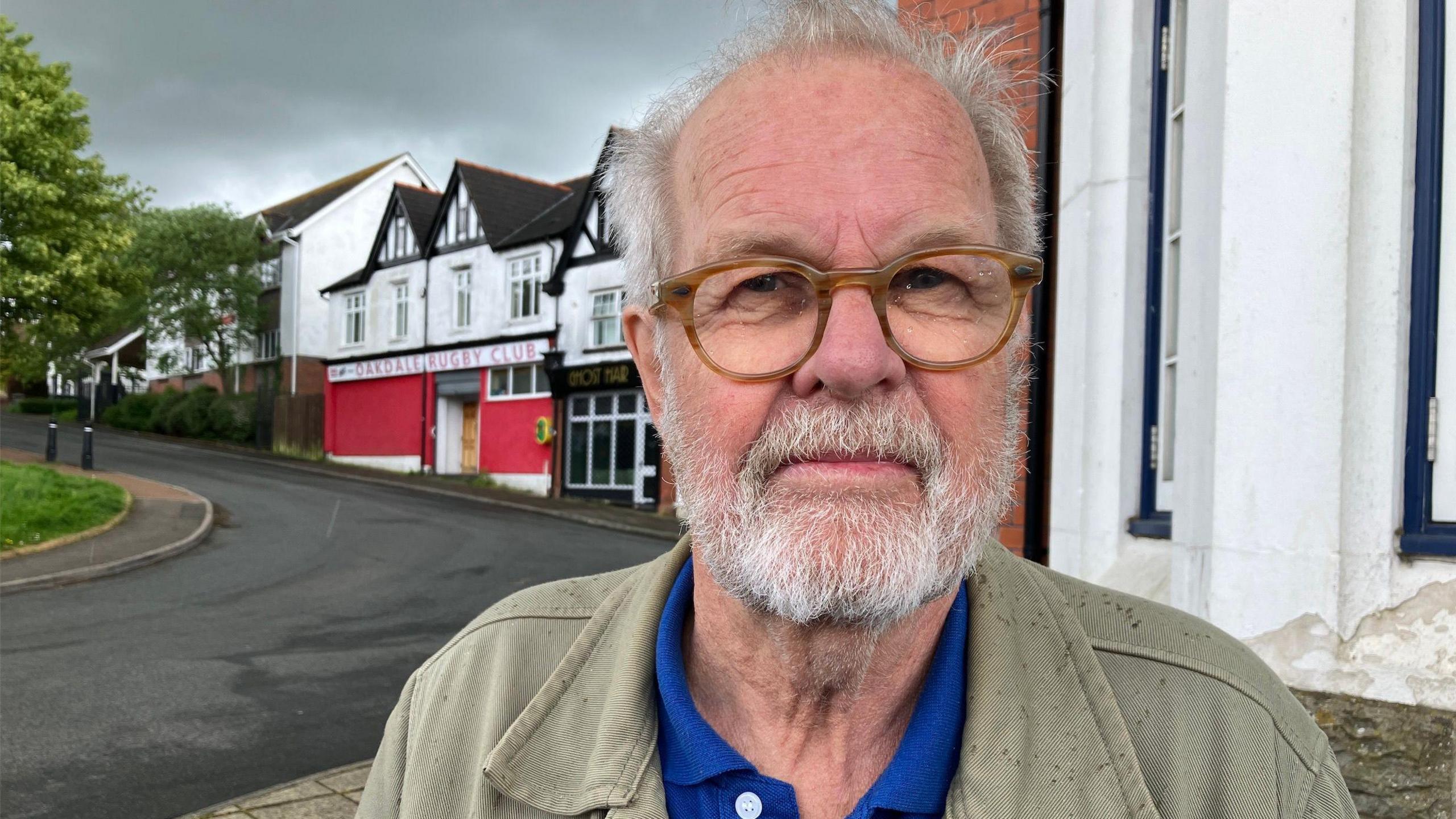 Photo of Kjell-Åke Andersson who has grey hair and a grey beard. He's  wearing brown rimmed glasses and wearing a blue polo tshirt and faded green jacket. Behind him over his right shoulder is a road and some terraced buildings including a rugby club entrance. The sky has grey clouds and there is also a patch of grass and part of a green tree visible to the left of the frame.