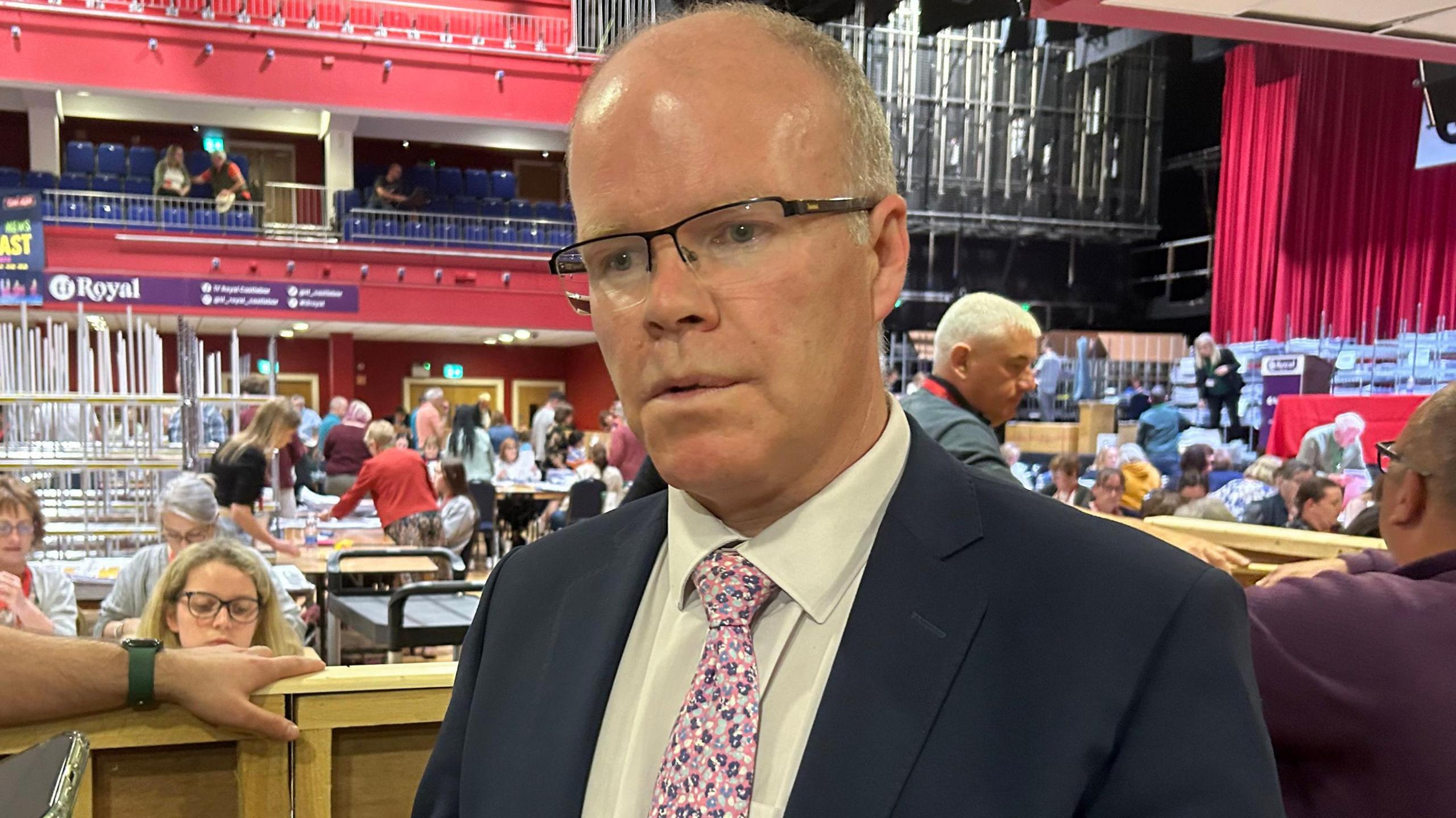 A man wearing black glasses, a navy suit, white shirt and pink Tie stands in a busy theatre