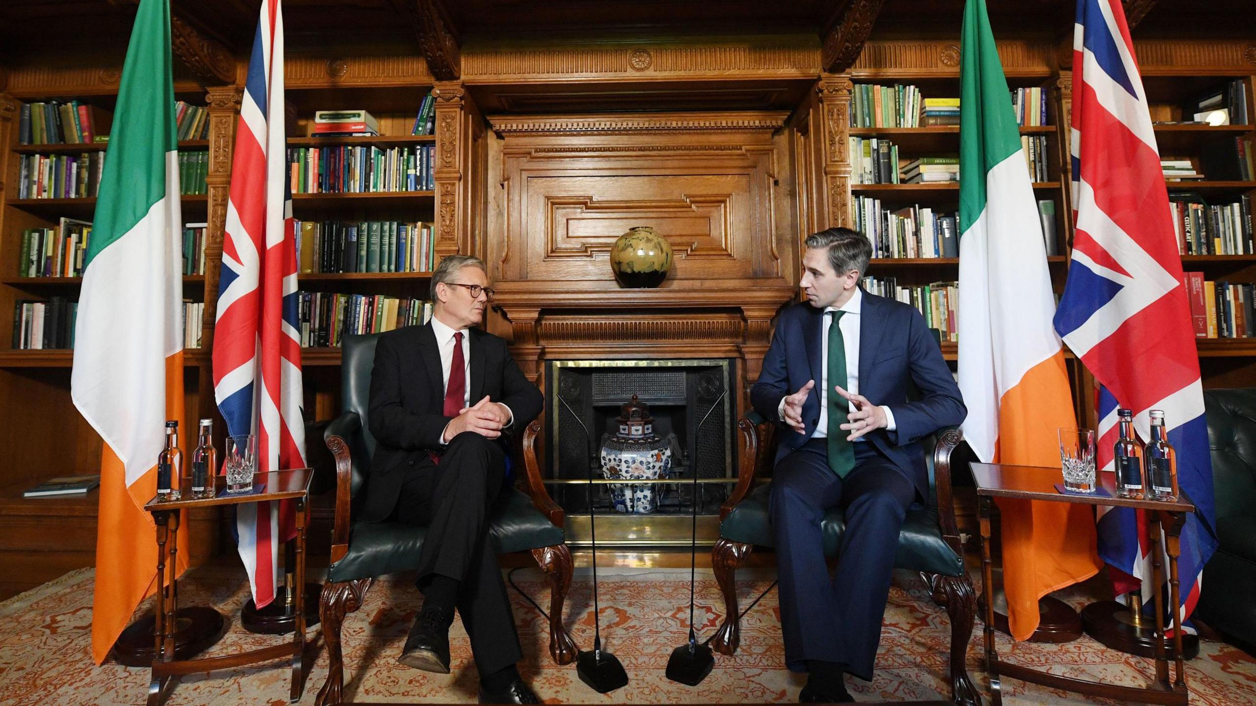 Sir Keir Starmer sitting with Simon Harris in library in Dublin's Farmleigh House, with British and Irish flags beside each of the two leaders