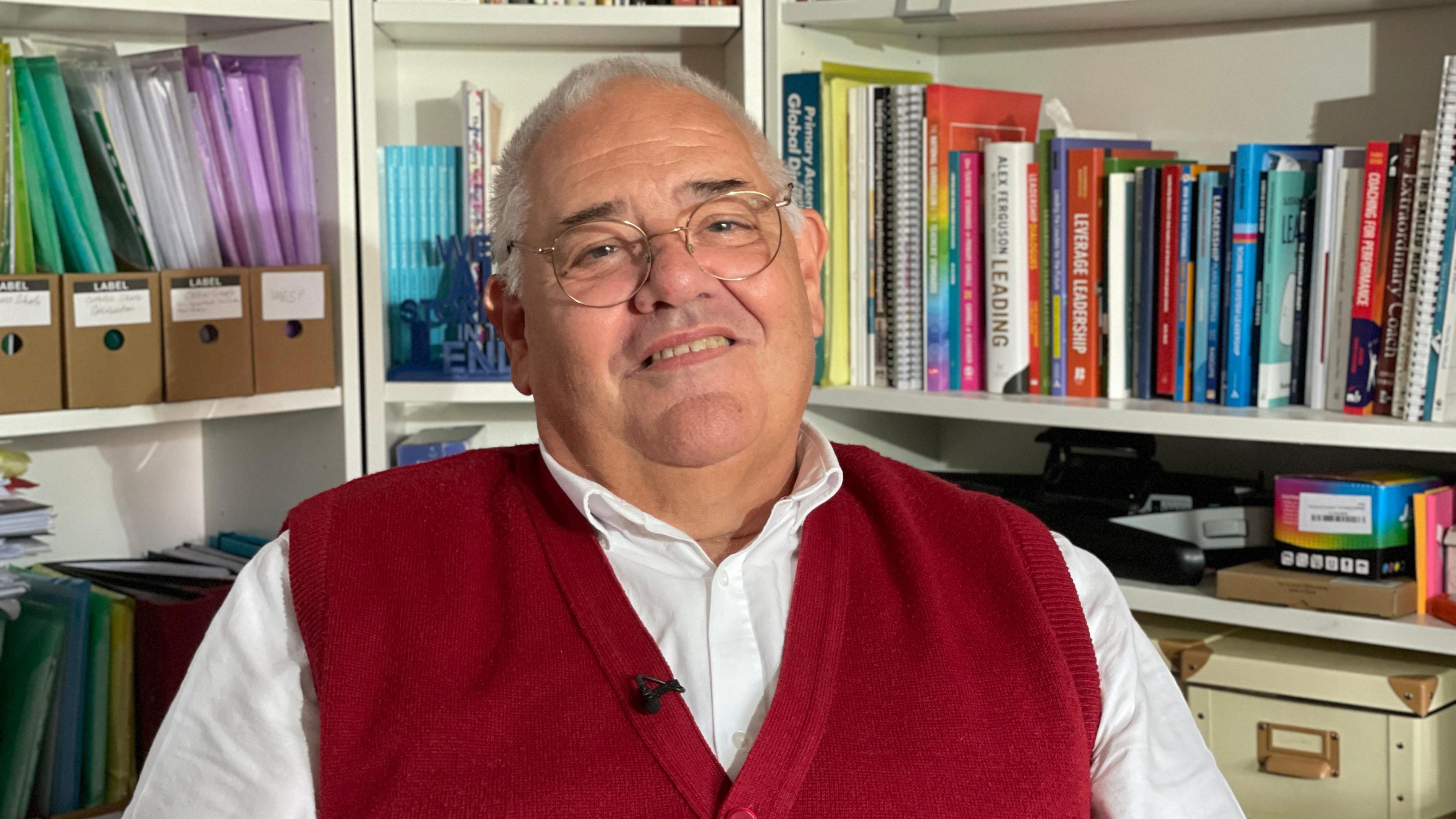 Bill Jerman - wearing glasses and red cardigan - looks into camera with bookshelves behind