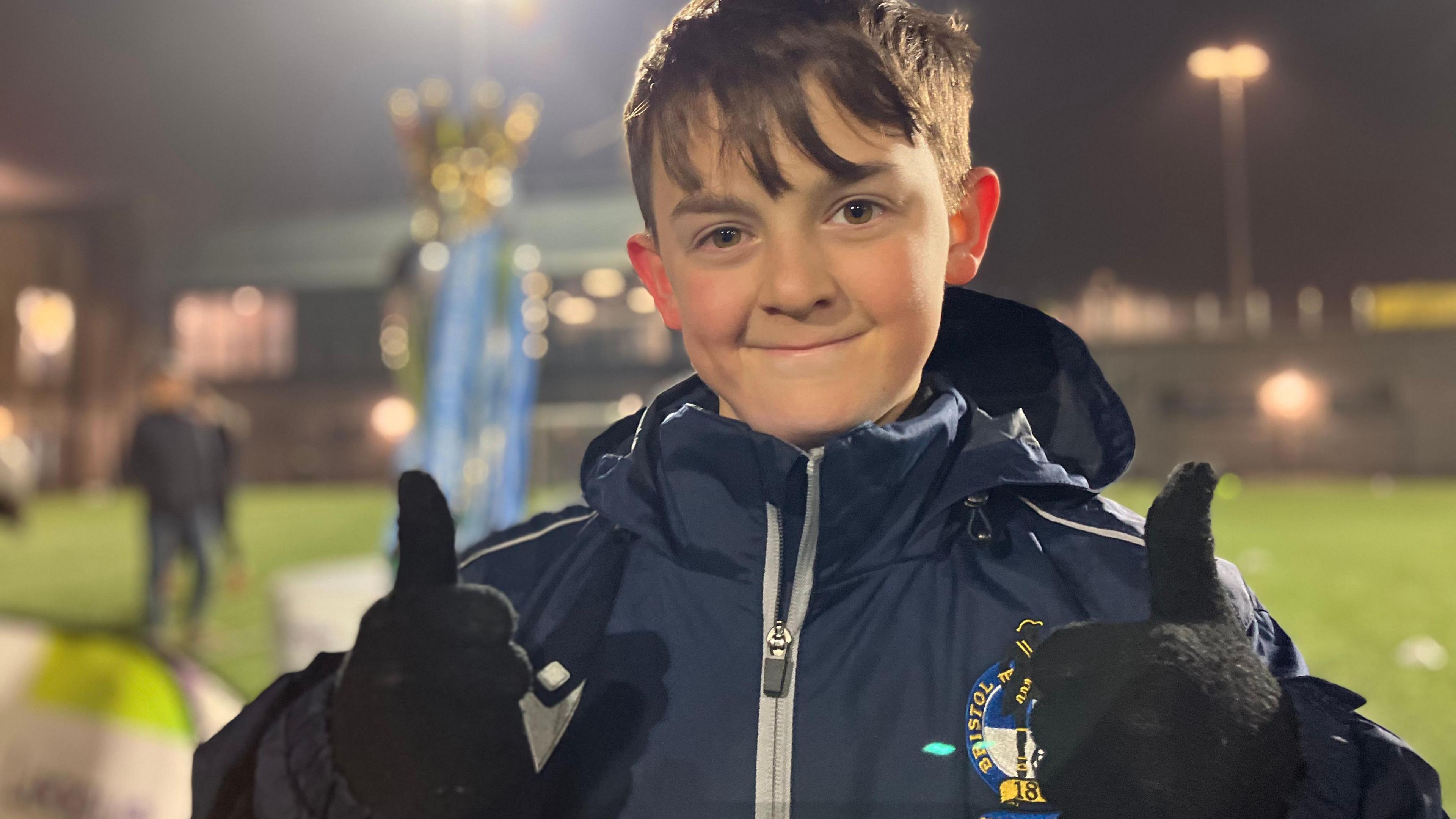 Cameron looking at the camera smiling, with both his thumbs pointing up and the Premier League trophy in the background.