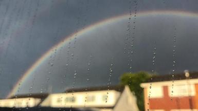 Looking through a window pane with raindrops running down it. Outside the window there are dark clouds and a rainbow