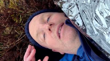 Glenn Campbell lying on the ground looking up at the camera, with a foil blanket over his body. He is wearing a blue woolen hat and blue clothing.