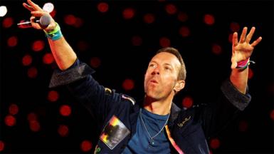 Chris Martin from Coldplay holding both hands up in the air, with a black microphone in his right hand. he is wearing a denim jacket and blue top underneath, with red spotlights behind him.