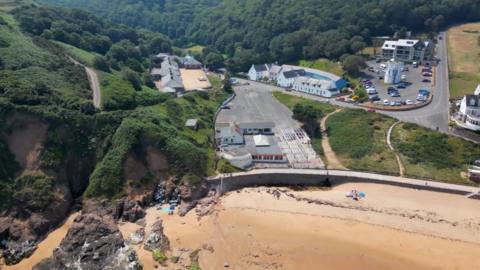 A drone photo of Greve De Lecq beach. At the bottom of the photo is the beach with browny-white sand, and on the left the sand turns into brown cliffs with green grass and hedges on top of them. There is also a car park and conrete.