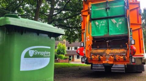 A green bin with a Shropshire Council logo on it and a bin lorry in the background