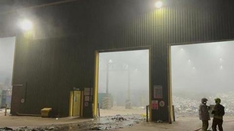 Firefighters stand at the entrance to a large smoke-filled metal shed with rubbish strewn across the floor