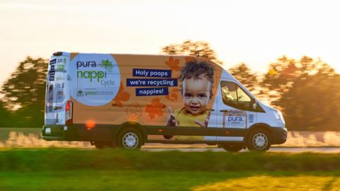 NappiCycle van seen driving by with trees in the background. The van is white with orange painting around the sides. On the side we're looking at, there's a picture of a baby with the text Holy poops we're recylcing nappies written in blue boxes in a white font.