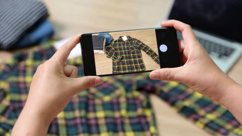 A person's hands holding a phone showing a photo of a checked shirt, which you can see is under the phone lay on a desk, looking less in focus