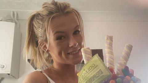 A young blonde woman with her hair tied in a ponytail. She is smiling at the camera and holding a cake covered in Marshmallows, chocolate sweets and a piece of yellow paper which reads "Golden Ticket".