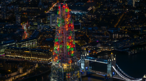 Shot from a helicopter at night, green, red and and orange lights shine in the top of the Shard with Tower Bridge in the background