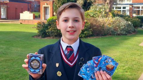 A boy wearing a school uniform holds a pack of cards in one hand and several playing cards in the other as he poses with them while looking at the camera. He is standing on a green lawn with houses behind him. It is a sunny day.