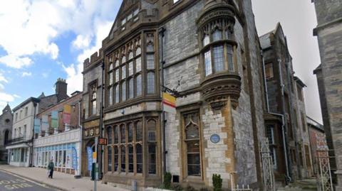 The front of the Dorset Museum on the sloping hill of High West Street - a stone building with large gothic-style bay windows with leaded glass on the ground and first floor and a crenellated parapet with a pitched roof