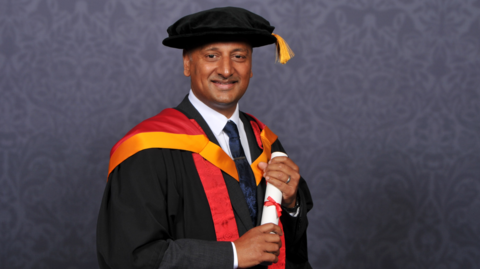 Mr Patel is in traditional university attire, wearing a gown and cap, and holding a scroll of paper. 
