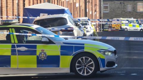 A police car parked on Elm Street, Stanley. A police cordon is visible at several points along the street, A blue tent has been put up outside a house. Other police vehicles are parked in the background.