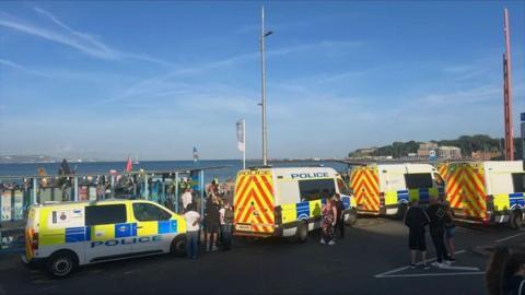 Line of police vans along seafront with crowds seen on the beach