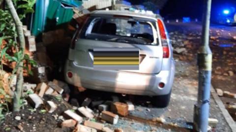 A silver car crashed with its front end into a brick wall. Surrounding the car are lots of red scattered bricks and there is also a green storage container that has been damaged