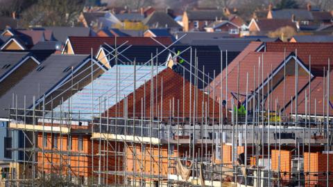 Scaffolding around two buildings on an under-construction housing estate. Two workmen in hi-vis jackets are working on the buildings.