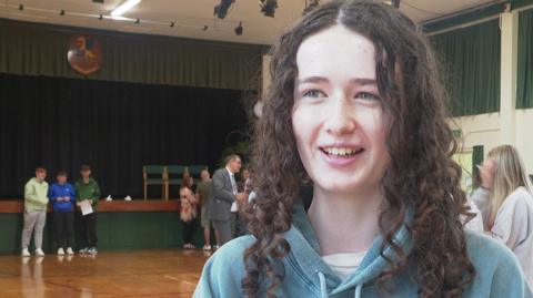 Grace, a Down High pupil has brown curly hair and wears a blue hoody. She smiles at the camera in her school assembly hall