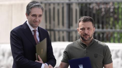 Simon Harris and Volodymyr Zelensky shaking hands and holding folders. 