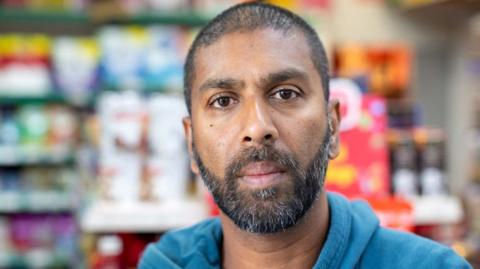 Shopkeeper Chanaka Balasuriya looks into the camera in his store, he is wearing a blue hoodie