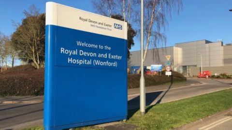 Exterior of Royal Devon and Exeter Hospital in the background and a blue and white sign with the name of the hospital and NHS logo in the forefront