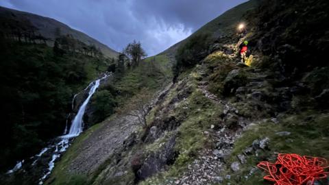 Taylorgill Force waterfall, Seathwaite, which shows a mountain rescue team member wearing a green helmet and another with a light on a helmet. A pile of orange rope is in the foreground.