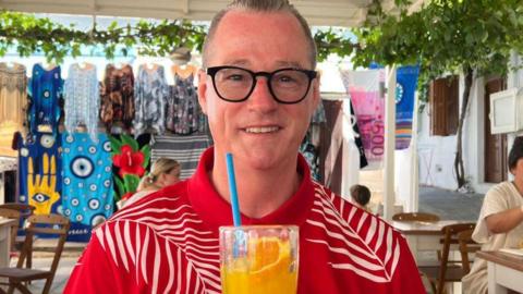 Man with short greying hair, smiling, looking at camera, wearing dark-rimmed glasses, wearing a red shirt with white pattern, holding a glass with orange liquid inside and an orange slice and blue straw, in what appears to be a cafe setting abroad, with bright towels and tops hanging up in the background.  