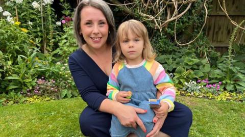 A smiling Danielle sitting on a lawn dressed in blue with Erin on her lap wearing a striped top