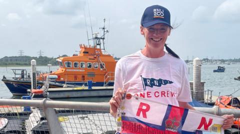 A woman wearing a blue hat and holding a flag which reads "RNLI". In the background there is an orange lifeboat on the water.