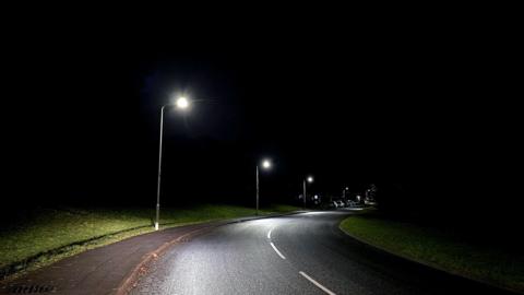 An LED lit empty road, there is a row of street lamps on the left hand side with a patch of grass behind them, the lamps are overlooking a section of road which curves round to the right of the image